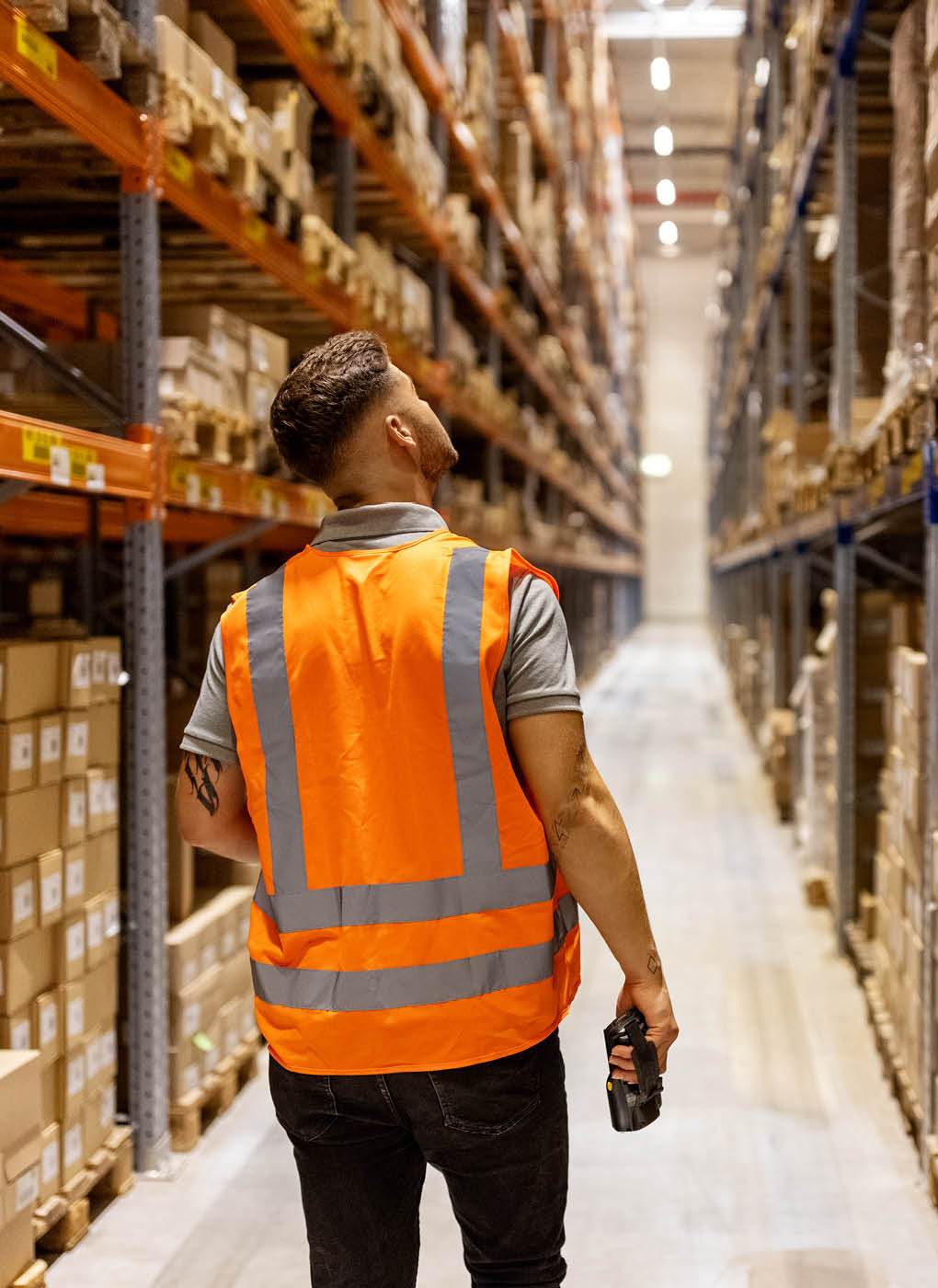 Rear view of a male worker in reflective clothing walking in the warehouse. Male employee working in large storage room verifying the stocks on rack.